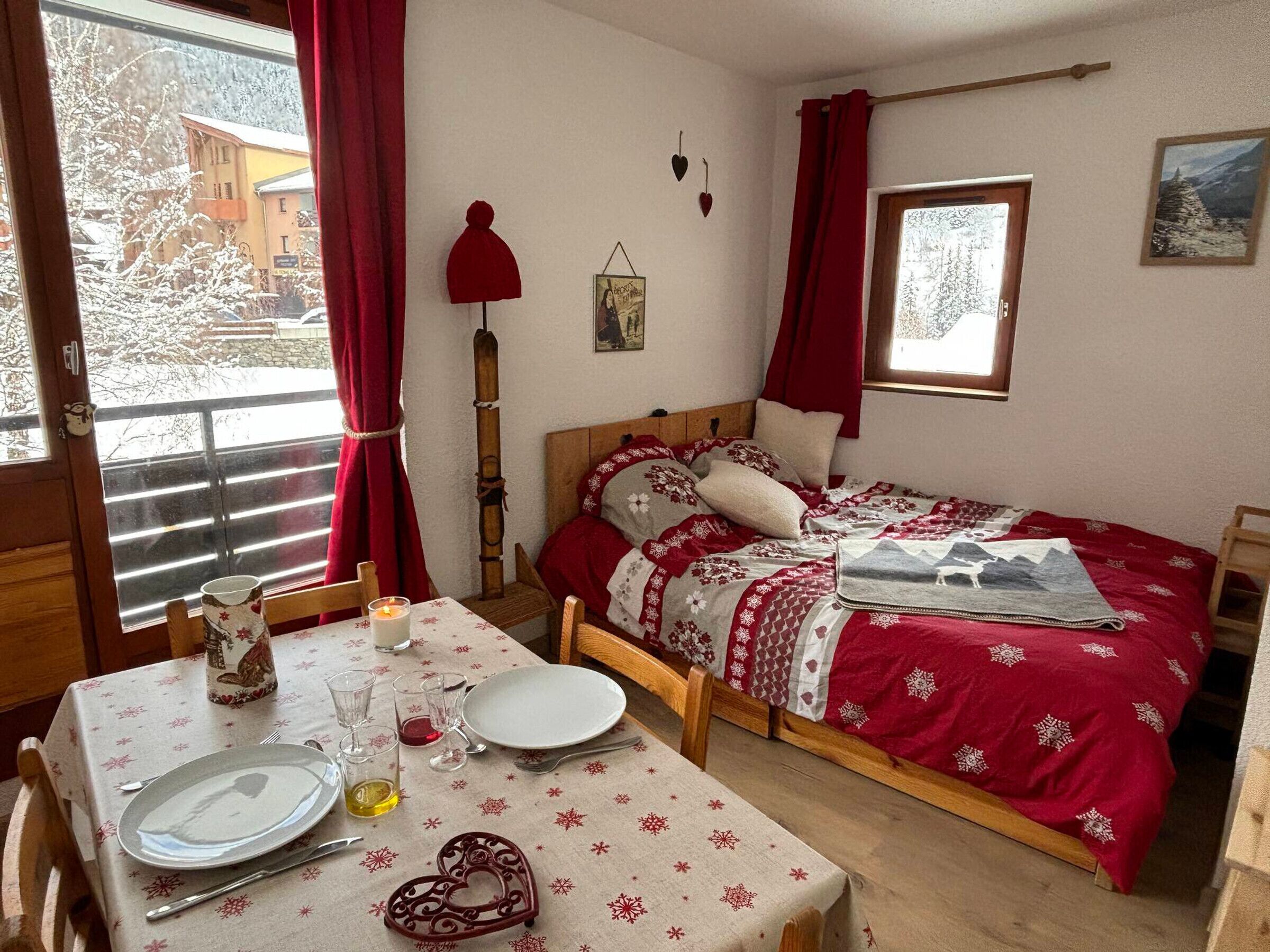 A cozy bedroom with a wooden bed, a dining table set with plates and glasses, and a window with a view of snowy trees.