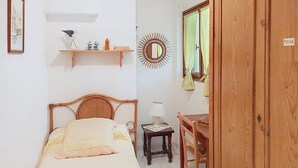 Un dormitorio con cama, una mesita de noche con lámpara, una silla, un armario de madera, un espejo y una estantería con objetos decorativos.