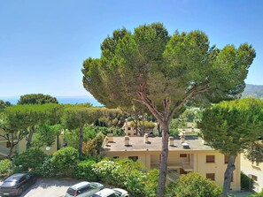 Una zona residenziale con un grande albero in primo piano, un edificio con balconi e una vista limpida sul mare sullo sfondo.
