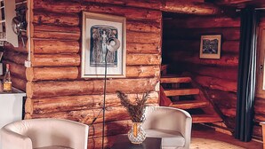 A log cabin interior with two white armchairs, a wooden floor, a woven ceiling light, and a framed artwork on the wall.