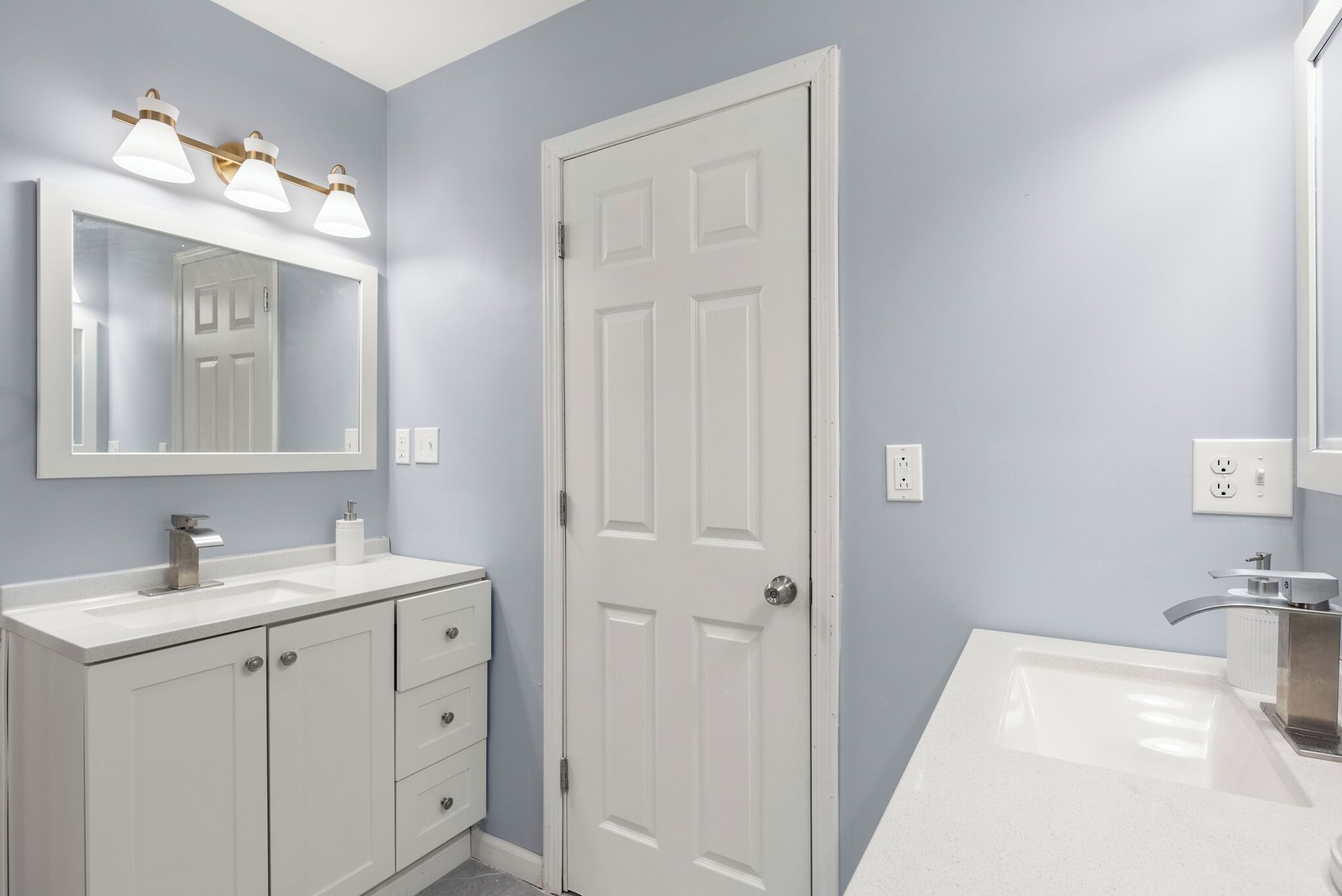 Une salle de bain avec un meuble-lavabo blanc, un double vasque et une porte surmontée d’un miroir.