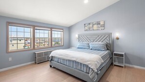 A bedroom with a large bed, a nightstand, a wall-mounted lamp, and a window with a view of buildings. - Bela Vista: Stunning Water Views, Hot Tub, Pets OK (Nags Head)