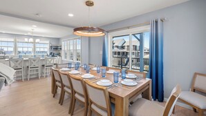 A dining area with a wooden table set for six, surrounded by chairs, with a view of a balcony and a cityscape through large windows. - Bela Vista: Stunning Water Views, Hot Tub, Pets OK (Nags Head)
