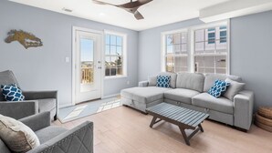 A modern living room with a grey sectional sofa, a small table, and a ceiling fan. - Bela Vista: Stunning Water Views, Hot Tub, Pets OK (Nags Head)