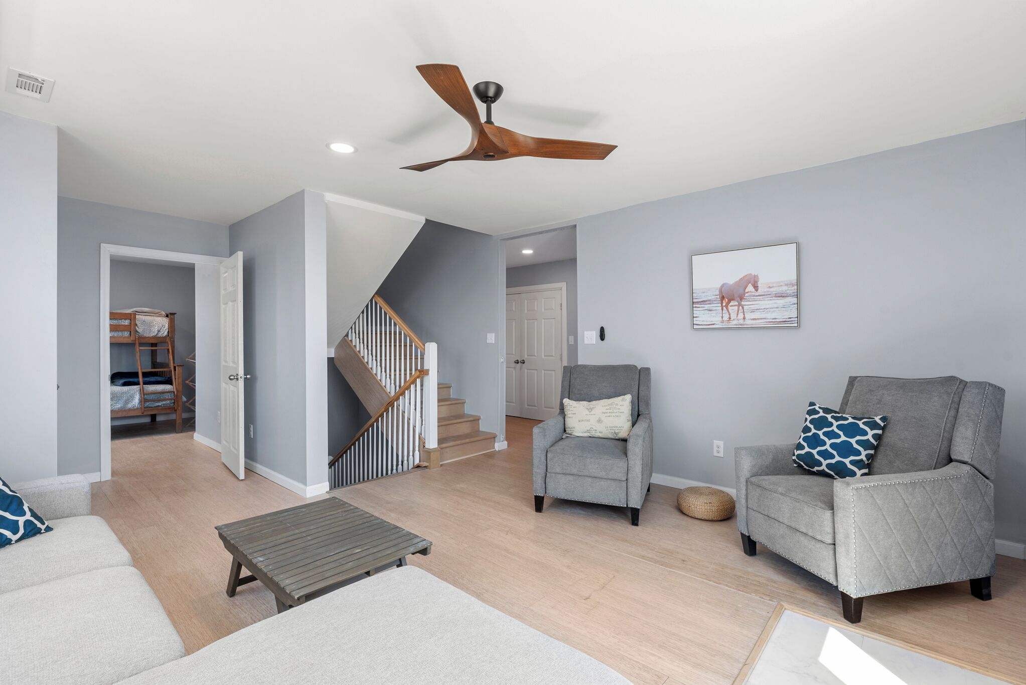 A spacious living room with a grey armchair, a sofa, and a wooden coffee table. A ceiling fan is visible, and a staircase leads to another room.
