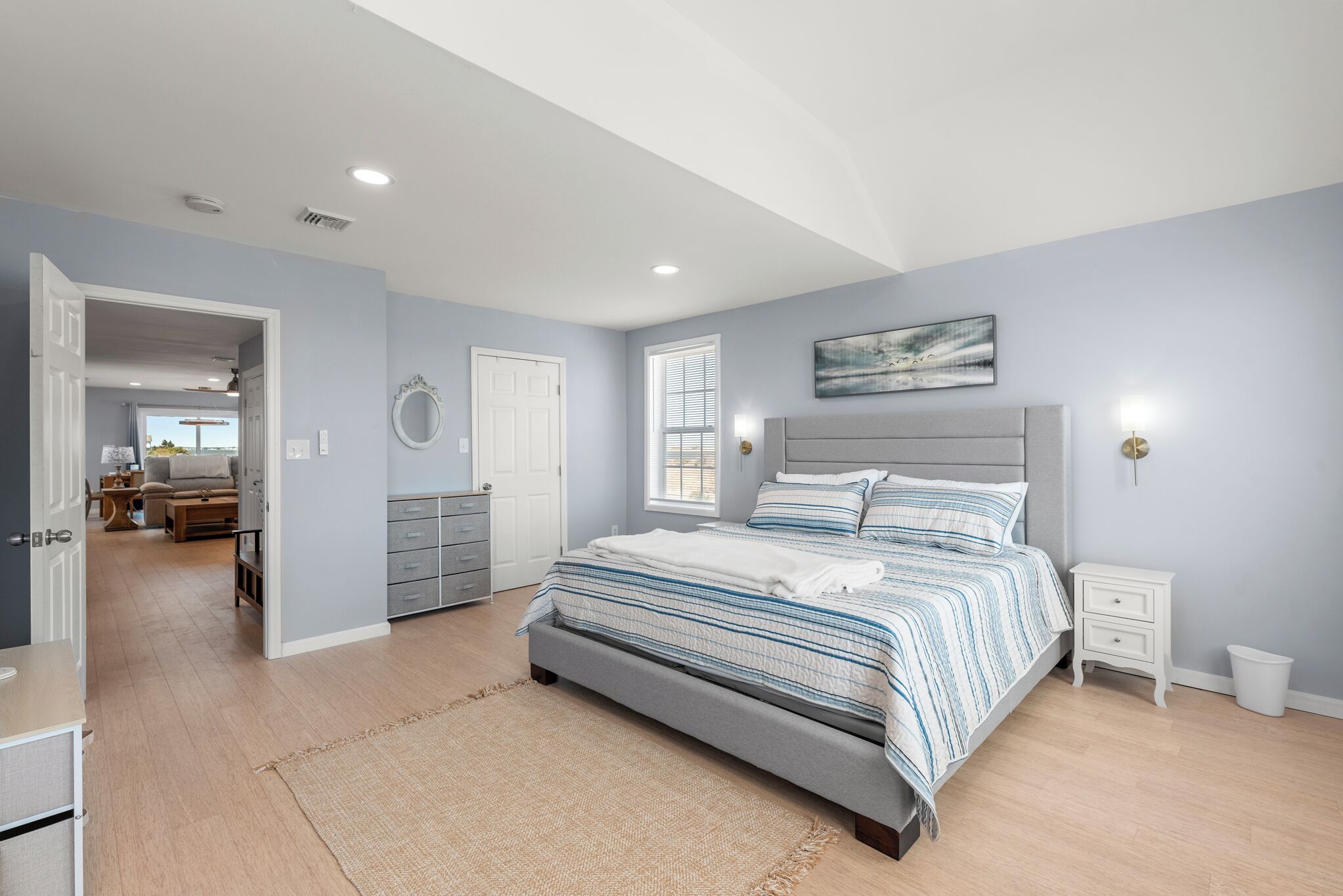A bedroom with a large bed, a bedside table, a nightstand, a wall-mounted light, and a door leading to another room.