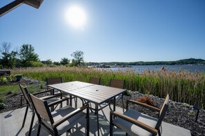 En udendørs terrasse med et bord og stole, med udsigt over en sø med både og en fjerntliggende bakke.