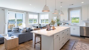 Een moderne keuken met witte kasten, een centraal eiland, hangende lampen en grote ramen met uitzicht op de omgeving.