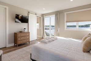 Ein Schlafzimmer mit Bett, Fernseher, Kommode und Blick auf ein Gewässer durch große Fenster.