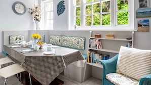 A dining area with a table set for four, a bookshelf, and a window with a view of greenery. - Stylish Dog-Friendly Cottage with Stunning Sea Views in Mousehole, secure garden (Mousehole)