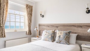 A bedroom with a large bed, a wooden headboard, a nightstand with a lamp, and a view of the sea through a window. - Stylish Dog-Friendly Cottage with Stunning Sea Views in Mousehole, secure garden (Mousehole)