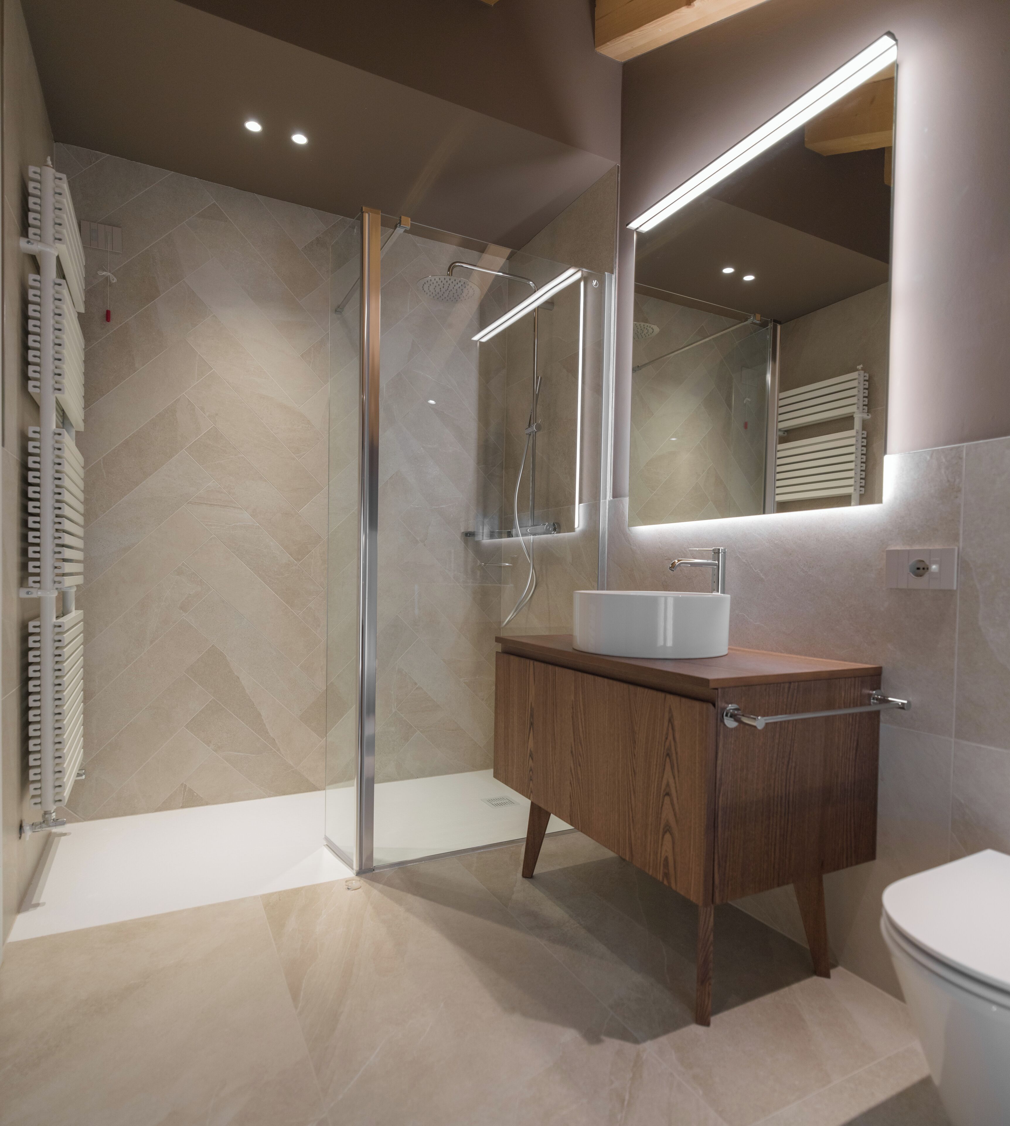 Un baño moderno con cabina de ducha de vidrio, tocador de madera con lavamanos blanco y un espejo grande sobre el tocador.