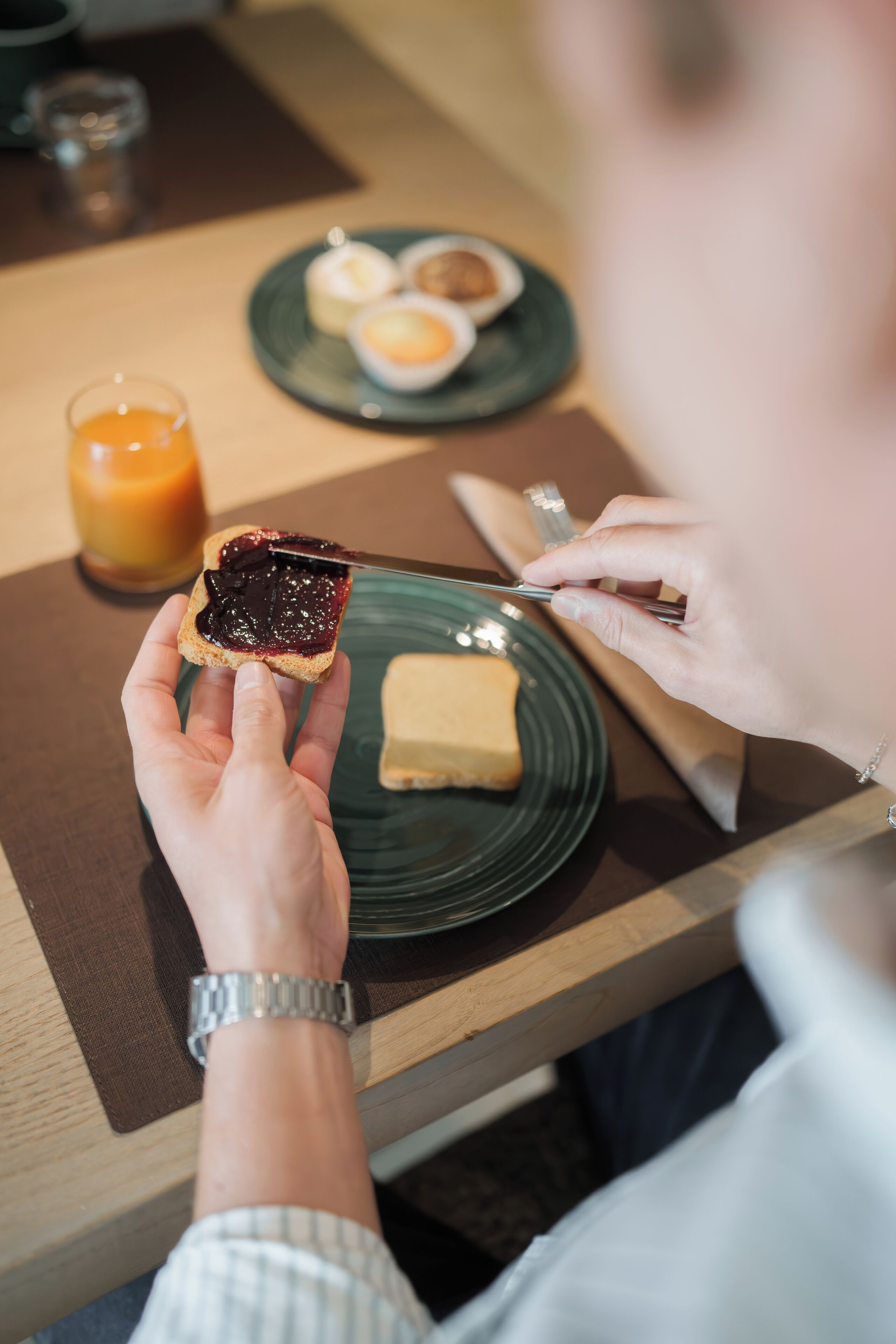 A person holding a slice of toast with jam, a knife, and a plate with a piece of butter.