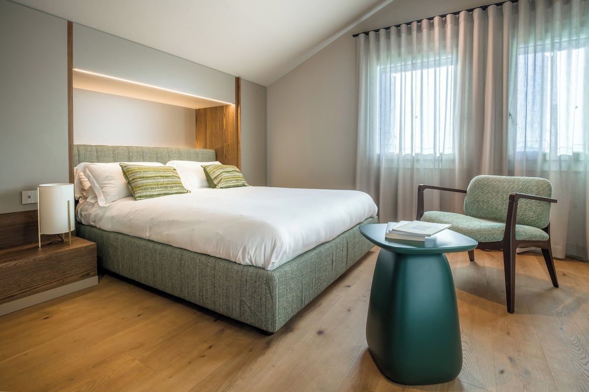 A modern hotel room with a large bed, a green upholstered chair, a small round table, and a wooden floor.