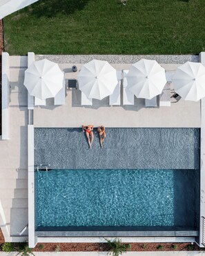 Vista aérea de una piscina con dos personas relajándose bajo sombrillas blancas, rodeadas de sillas de descanso y un césped cuidado.