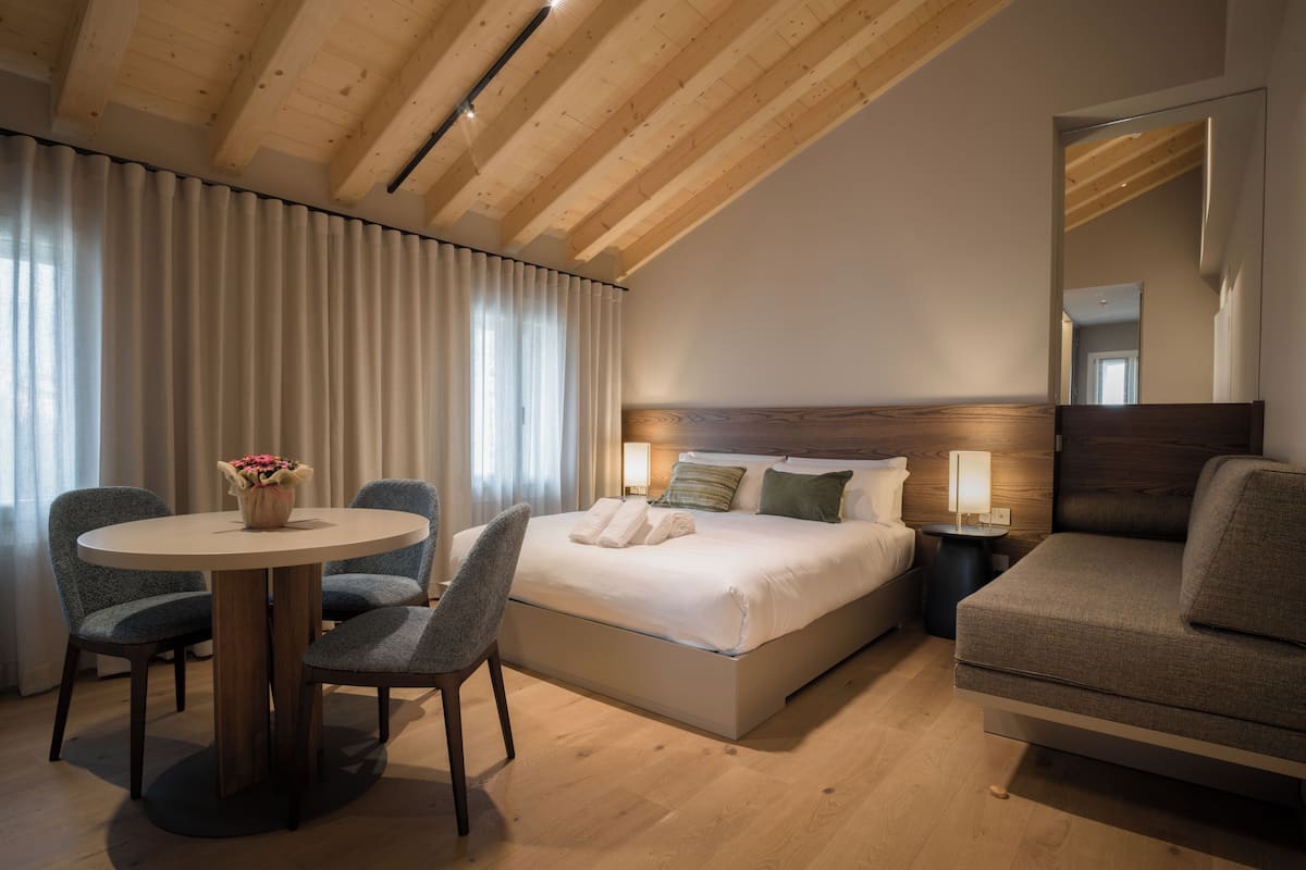 A hotel room with a wooden ceiling, a bed with white linens, a round table with two chairs, and a sofa.