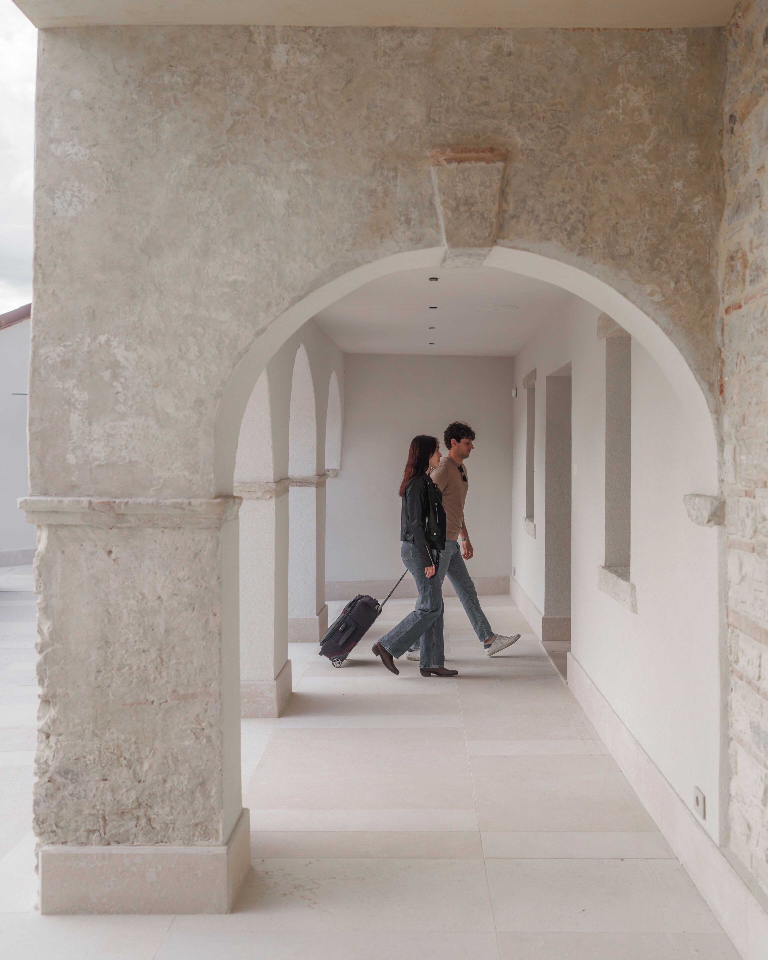 A corridor with a stone archway, a person pulling a suitcase, and another person walking beside them.