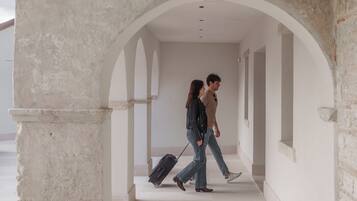 A corridor with a stone archway, a person pulling a suitcase, and another person walking beside them.