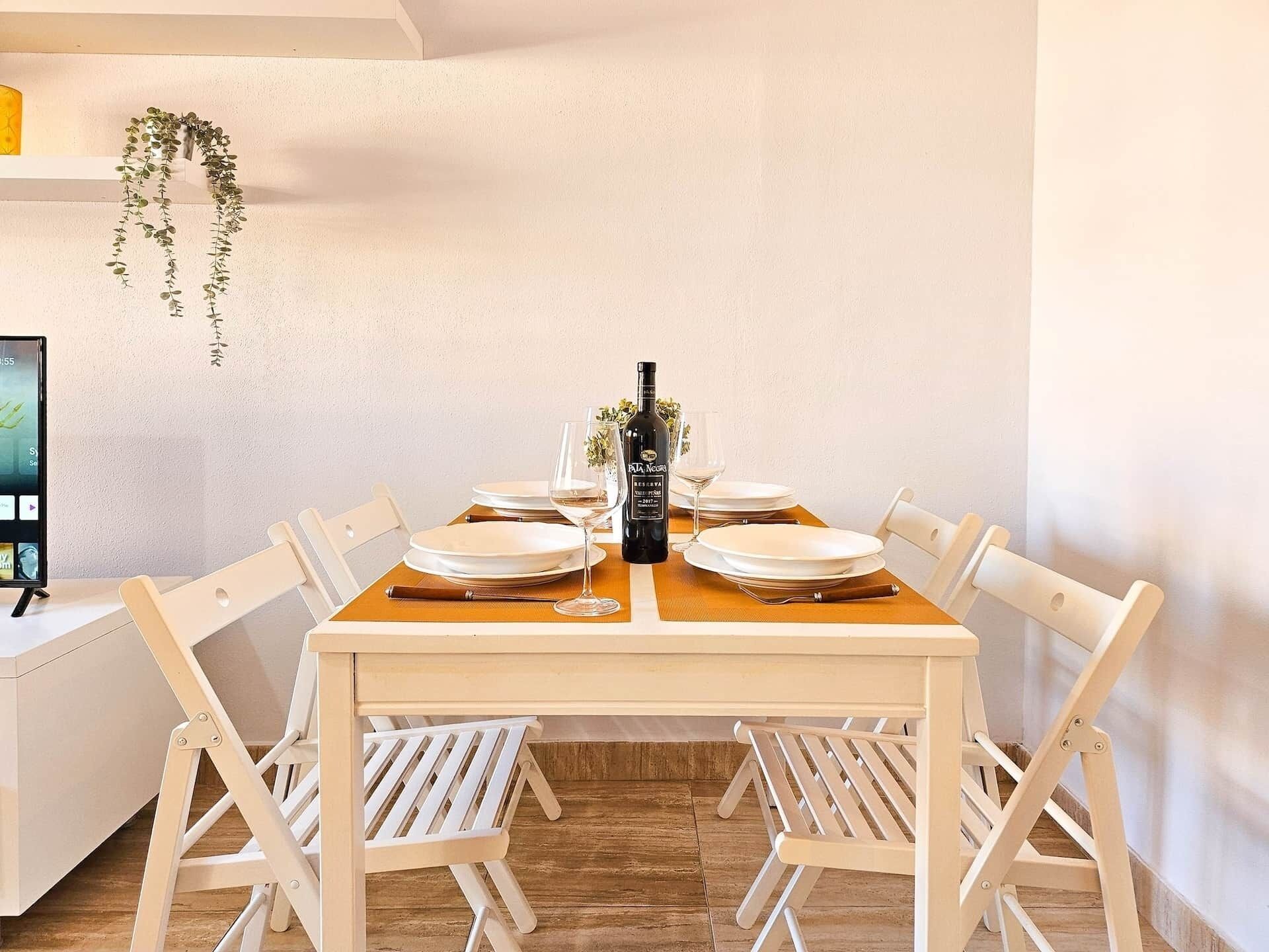 Een eettafel gedekt met borden, glazen en een fles, omringd door witte stoelen in een kamer met een houten vloer en een witte muur.