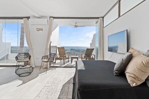 Habitación de hotel moderna con balcón, una cama, dos sillas, televisión y vistas a la ciudad.