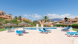 Un área de piscina con sillones de descanso y sombrillas, rodeada de edificios y palmeras.