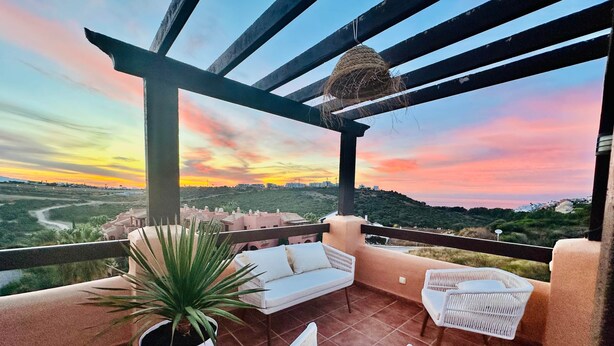 Un balcón con mobiliario de mimbre, una planta en maceta y vistas a un paisaje al atardecer.