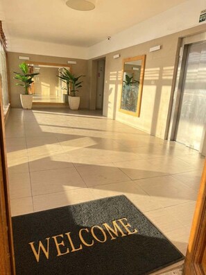 A hotel lobby with a reception desk, potted plants, and a welcome mat.