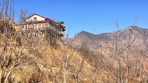 A building with a red roof and multiple floors, surrounded by dry, leafless trees and mountains in the background. - Apple velley homestay shanghar  (Sainj)
