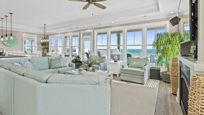 Een ruime woonkamer met een grote hoekbank, een plafondventilator en uitzicht op de oceaan door grote ramen.