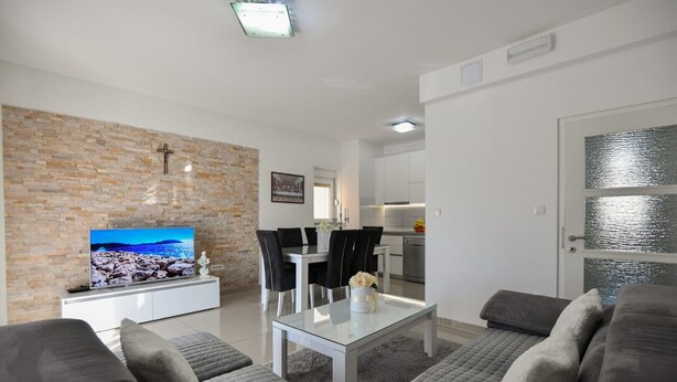 A modern living room with a stone wall, a flat-screen TV, a dining area with a glass table, and a grey sofa. - Apartment Dijana with private pool (Donji Prolozac)