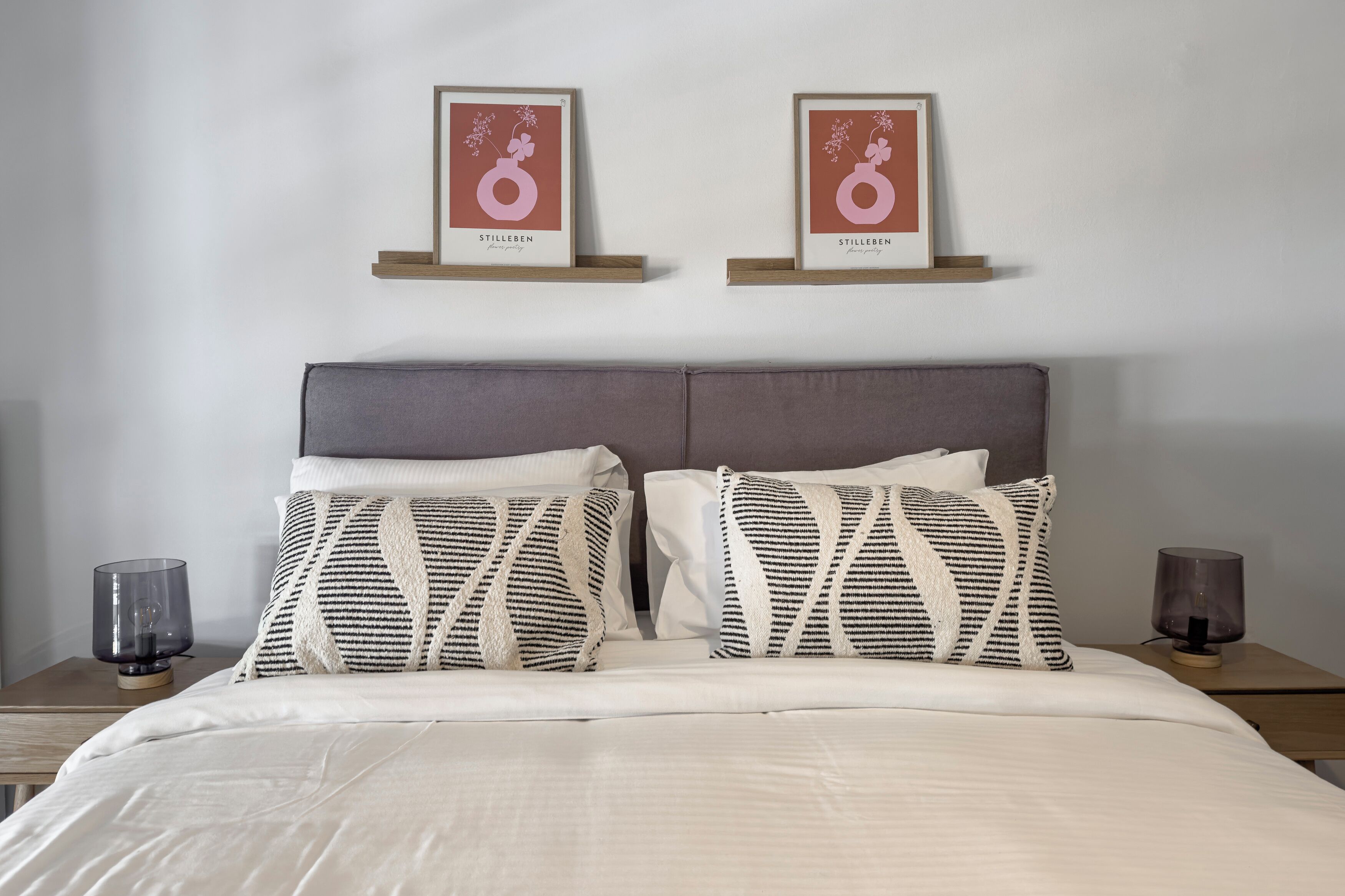A neatly made bed with striped pillows, a grey headboard, and two framed pictures on the wall.