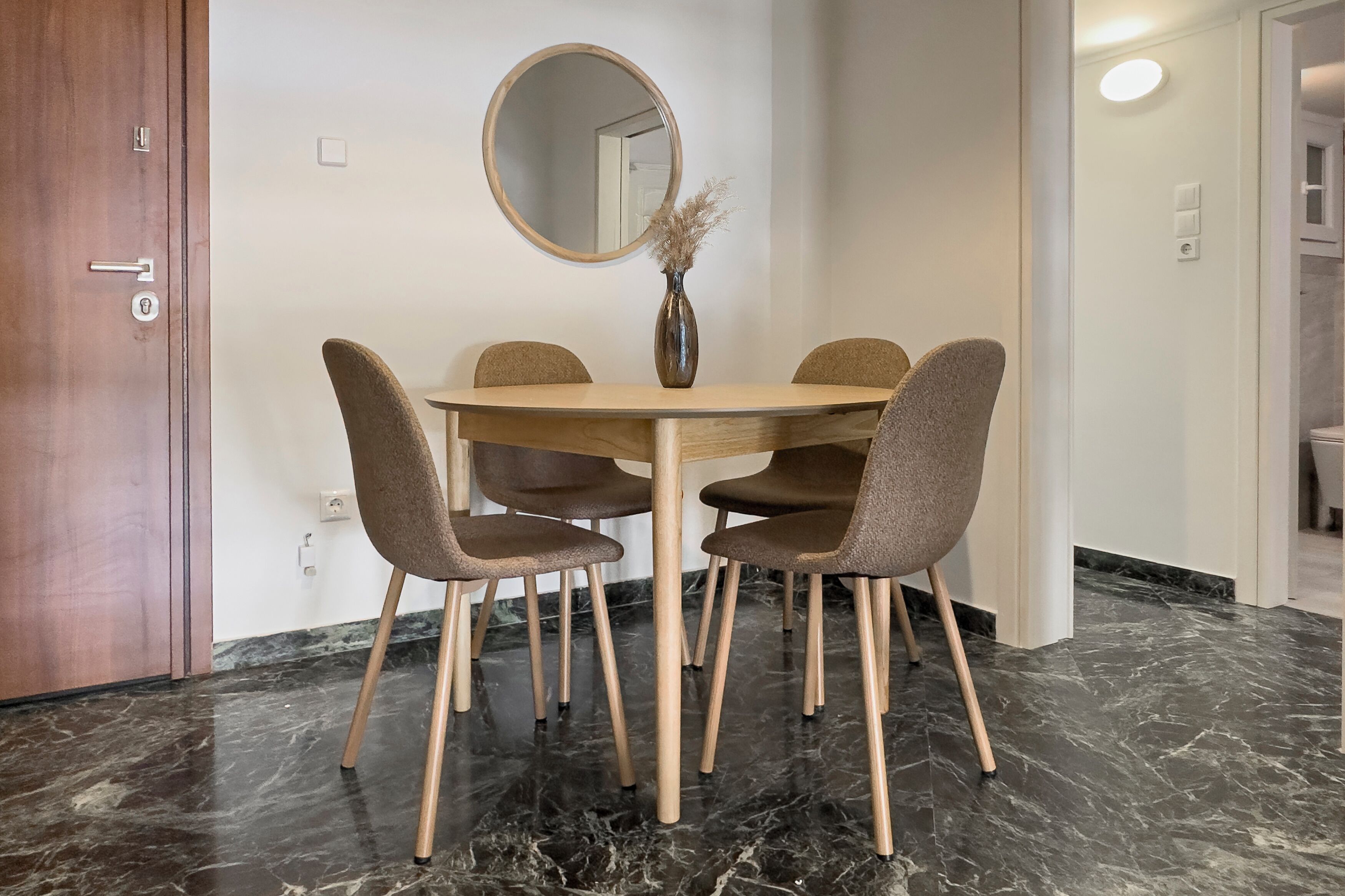 Spiseområde med et rundt bord, fire stole, en vase med tørrede planter og et rundt spejl på væggen.