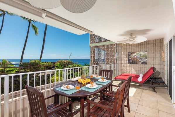 A balcony with a table set for two, chairs, a red lounge chair, and a view of the ocean.
