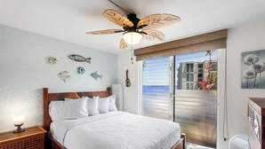 Ein Schlafzimmer mit einem Bett, einem Deckenventilator, einem Nachttisch und Blick auf das Meer durch eine Schiebetür.