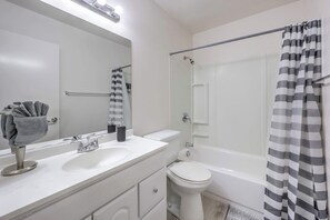 Un baño moderno con lavamanos blanco, espejo e inodoro. Hay una cortina de ducha y un perchero con toallas.