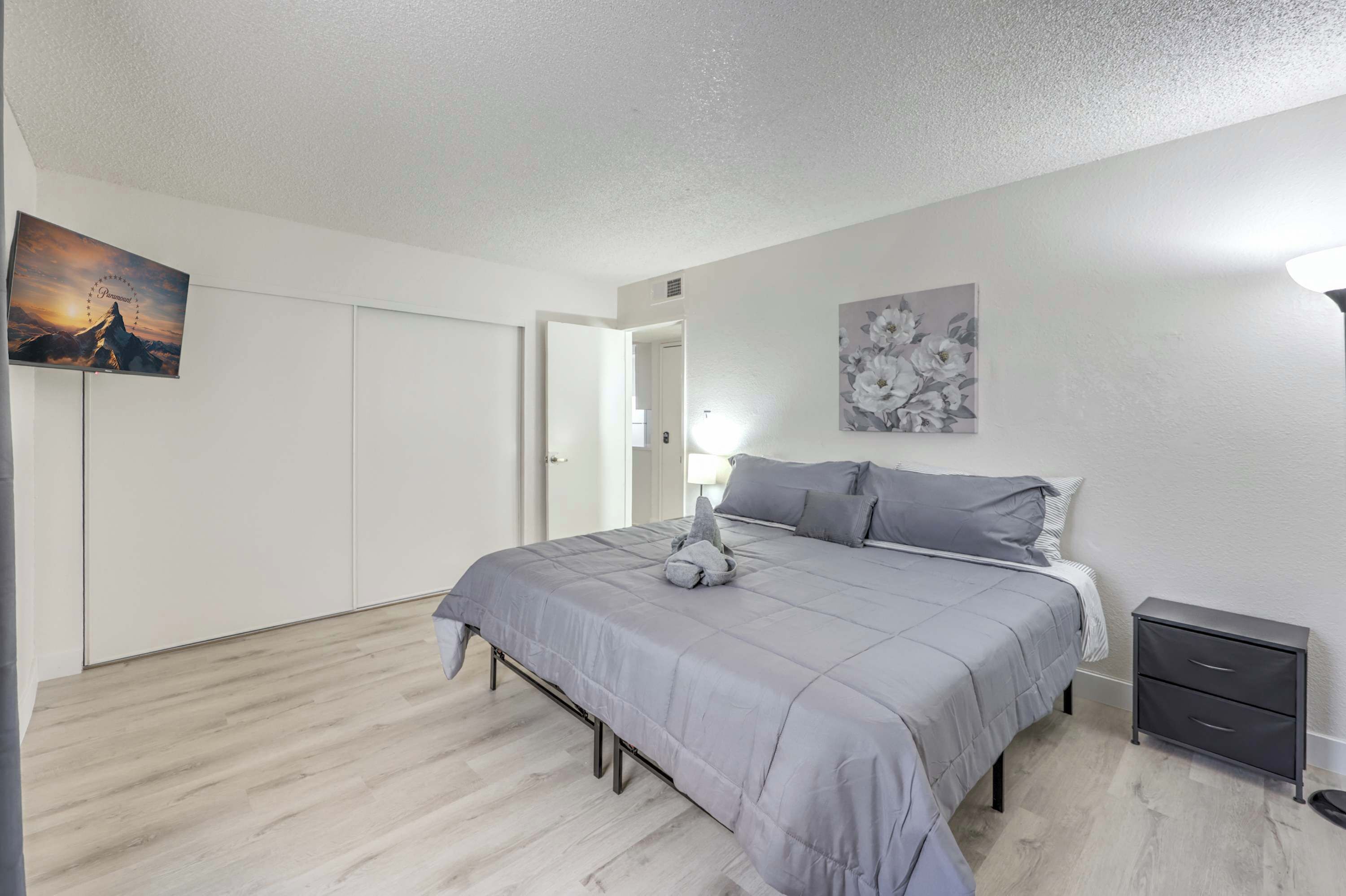 Um quarto com cama, mesa de cabeceira e televisão presa na parede.