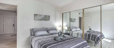 A bedroom with a large mirror, a bed with striped bedding, a bedside table with a lamp, and a marble artwork on the wall.