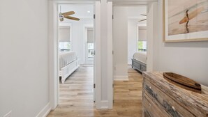 Ein Flur mit Holzboden, weißen Wänden und einem Deckenventilator. Ein Bett mit weißem Kopfteil und eine Kommode mit Holdecke sind ebenfalls vorhanden.