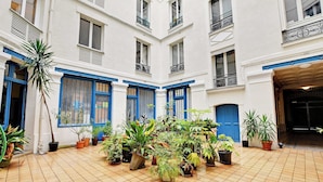 A courtyard with multiple potted plants arranged in a central area, surrounded by white walls with blue accents and multiple windows. - Cozy Flat 1 BDR / 2P - Marais District (Paris)