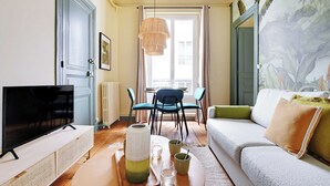 A living room with a television, a sofa, a dining table with chairs, and a large window with curtains. - Cozy Flat 1 BDR / 2P - Marais District (Paris)