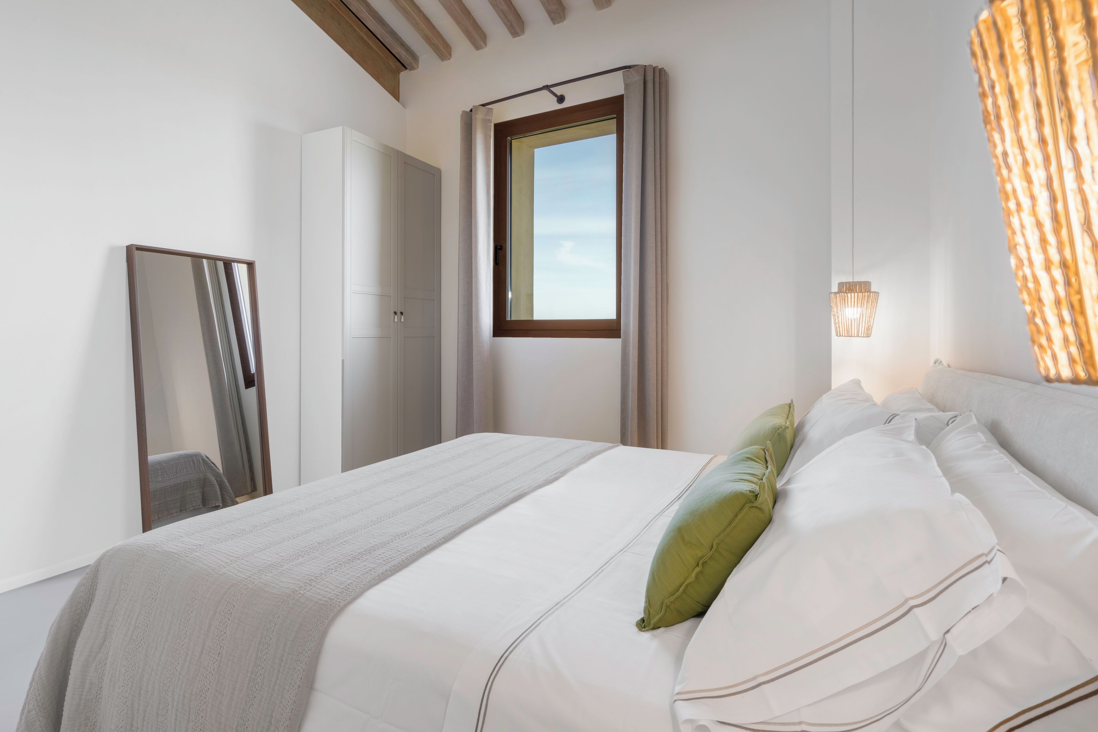 Habitación de hotel con cama, espejo, armario, ventana con cortinas y una lámpara de pared.