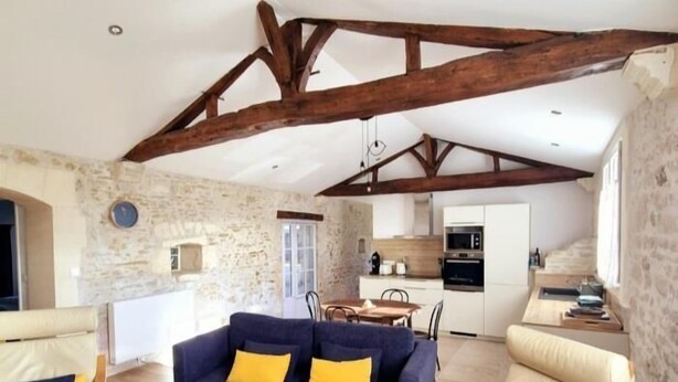 A spacious living area with a stone wall, wooden beams, a sofa, a chair, a wooden coffee table, and a kitchen in the background. - Old 18th century stable for 4 people (VERGEROUX)