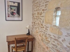 Una habitación con pared de piedra, un escritorio de madera, una silla, un cuadro enmarcado y una ventana.