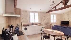 A kitchen with a wooden table and chairs, a stove, and a sink. There is a stone wall and a blue sofa in the living area. - Old 18th century stable for 4 people (VERGEROUX)