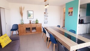 Un comedor moderno con una mesa y sillas de madera, un sofá con un cojín amarillo, y una cocina visible a través de una puerta abierta.