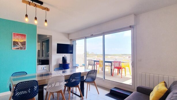 Una sala de estar moderna con un sofá, una mesa de comedor con sillas, un televisor y un balcón con sillas de colores.