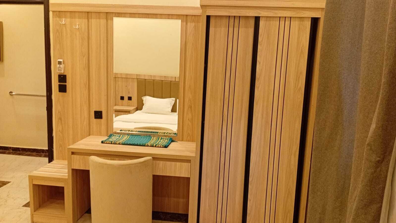 A compact hotel room with a wooden desk, a small chair, a bed with white bedding, and a large mirror.