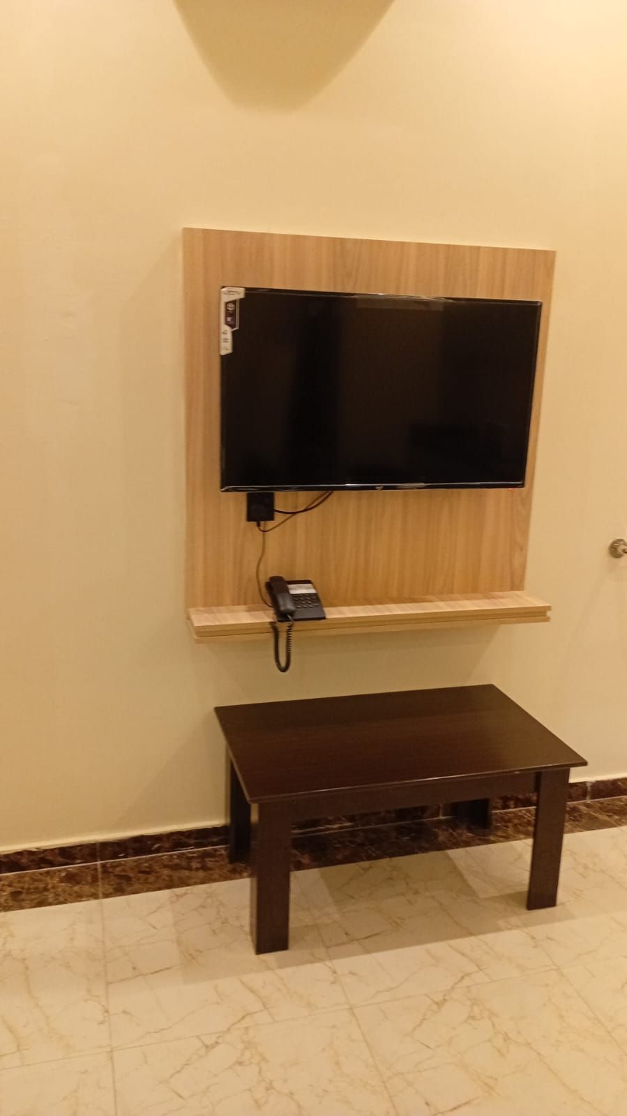A wall-mounted TV with a phone on a wooden shelf, a small table below, and a tiled floor.
