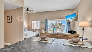 A hotel room with a dining area, a sofa, a TV, and a balcony with a view of the beach.
