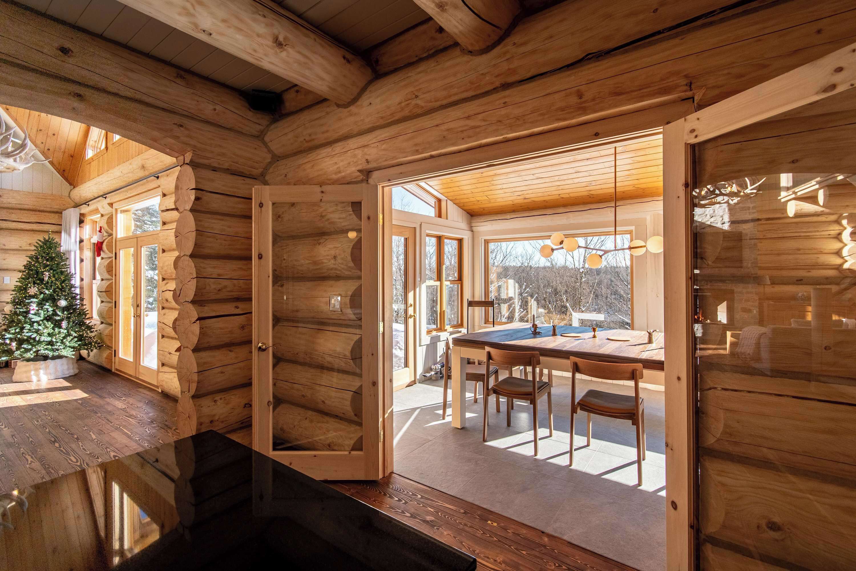 Interior de una cabaña de madera con comedor, un árbol de Navidad y grandes ventanales.